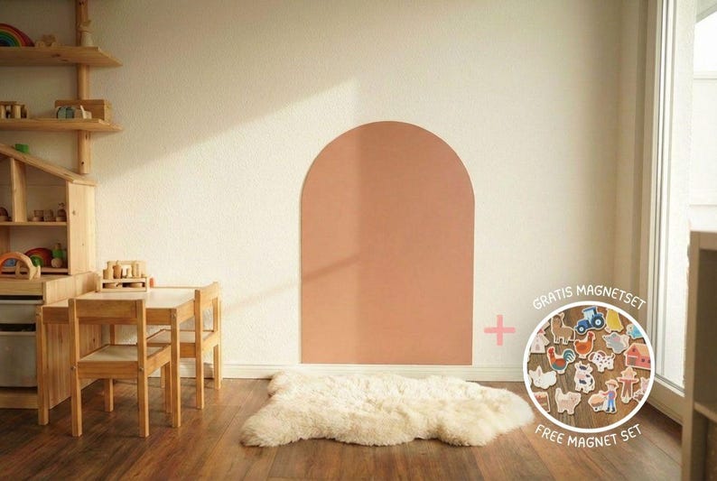 Ein Kinderspielzimmer mit Holzmöbeln, darunter ein Tisch und Stühle, und ein Regal mit Spielzeug. Ein großes, bogenförmiges Wandelement ist in einem gedämpften Korallenfarbton gestrichen. Ein weißer Schaffellteppich liegt auf dem Holzboden.