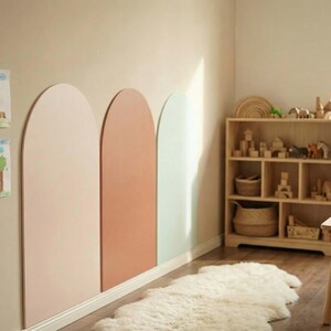 May include: Three large, vertical, arch-shaped wall panels in muted shades of light pink, terracotta, and pale mint green are mounted side-by-side on a beige wall, accompanied by children's drawings, a wooden shelving unit, and a small table.