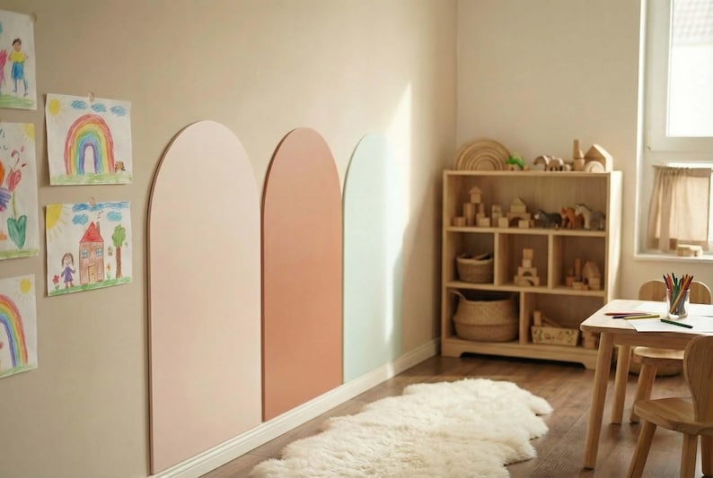 May include: Three large, vertical, arch-shaped wall panels in muted shades of light pink, terracotta, and pale mint green are mounted side-by-side on a beige wall, accompanied by children's drawings, a wooden shelving unit, and a small table.