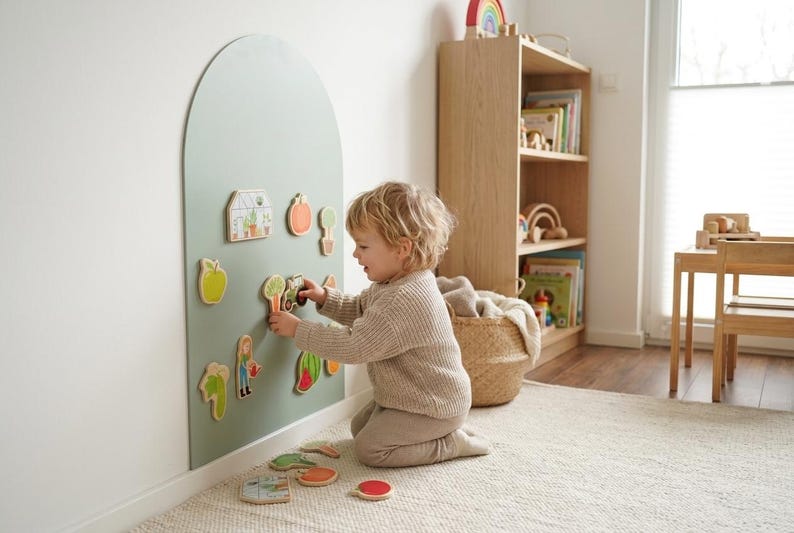 May include: A sage green arched wall-mounted magnetic board featuring various wooden garden-themed shapes including a greenhouse, vegetables, and a person with a watering can, with additional loose pieces resting on the floor.