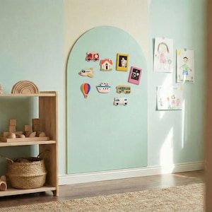 May include: A light green arched magnetic wall board featuring various colorful vehicle and frame magnets, positioned next to a wooden shelving unit filled with wooden blocks, rainbow stackers, and woven baskets in a room with light green walls.