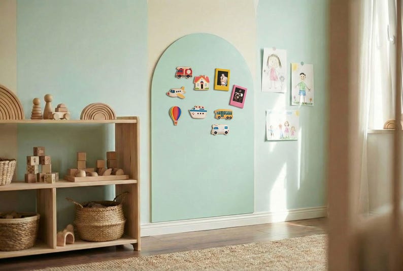 May include: A light green arched magnetic wall board featuring various colorful vehicle and frame magnets, positioned next to a wooden shelving unit filled with wooden blocks, rainbow stackers, and woven baskets in a room with light green walls.