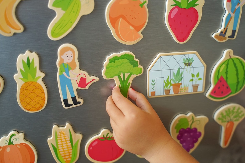 Ein Kind hängt die Obst und Gemüse Magnete auf den Kühlschrank. Es hält die Brokkoli Figur in der Hand.