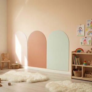 May include: A room interior featuring three large, arched wall decals in light pink, terracotta, and sage green. The space includes a light wood bookshelf filled with toys, a small wooden chair, and two white faux fur rugs on the floor.