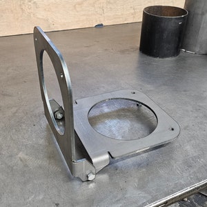 May include: A metal bracket with a circular cutout, designed to be mounted to a surface. The bracket is hinged and can be folded down.