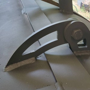 May include: Close-up of a black metal hinge with a curved arm and a bolt. The hinge is attached to a metal surface.