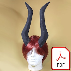 Puede incluir: Un par de cuernos negros grandes y curvos unidos a una peluca roja sobre una cabeza de maniquí blanca. Los cuernos tienen textura y apuntan hacia arriba. Un icono PDF está en la esquina inferior derecha.