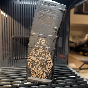 Può includere: Un caricatore per fucile in polimero nero con un'illustrazione incisa al laser di una figura barbuta in tunica che impugna un fucile. La parte superiore del caricatore riporta il testo 5.56 x 45, PMAG 30, GEN M3 e MAGPUL.