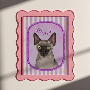 Op de afbeelding: Een ingelijste illustratie van een Sphynx-kat genaamd Elvira. De kat is grijs met groene ogen en is gecentreerd in een paarse ovaal met de naam Elvira. De achtergrond heeft paarse en witte strepen met gouden sterren. De lijst is roze.