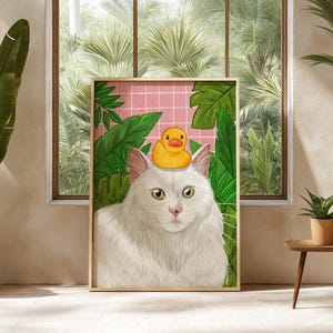 May include: A framed art print depicting a white cat wearing a yellow rubber duck on its head, set against a backdrop of green foliage and a pink grid. The artwork is displayed in a room with a window and plants.
