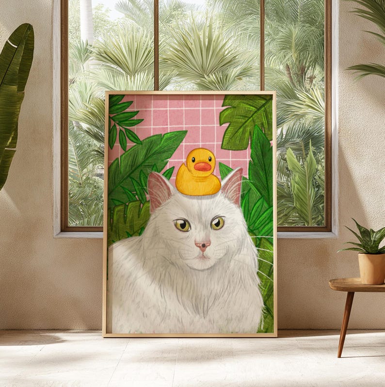 May include: A framed art print depicting a white cat wearing a yellow rubber duck on its head, set against a backdrop of green foliage and a pink grid. The artwork is displayed in a room with a window and plants.