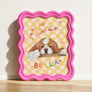 Op de afbeelding: Een ingelijste illustratie met een Cavalier King Charles Spaniel hond met bruin en wit haar, rustend op een houten richel. Het kunstwerk staat tegen een gele geruite achtergrond en is omlijst door een golvende roze rand. Tekst bevat "You are so loved" en de naam "BELLA".