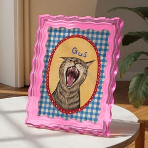 Op de afbeelding: Een roze fotolijst met golvende randen met een kattenportret. De kat gaapt, met de naam "Gus" erboven. De achtergrond heeft een blauw-wit geruit patroon.
