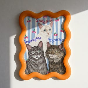 Op de afbeelding: Een ingelijst kunstwerk met drie katten met de woorden "Soda Pop", "Beauregard" en "Moonsug". Het kunstwerk heeft een blauw-wit gestreepte achtergrond met kersenillustraties en is ingekaderd in een golvend oranje frame.