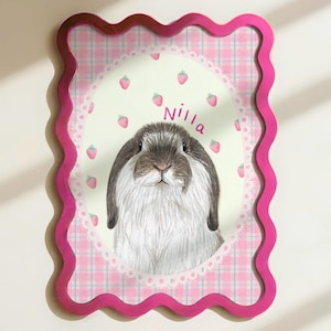 Peut inclure: Portrait encadré d'un lapin avec un bord festonné rose. L'œuvre présente un lapin au pelage gris et blanc, entouré de fraises et du nom "Nilla". L'arrière-plan présente un motif à carreaux roses.