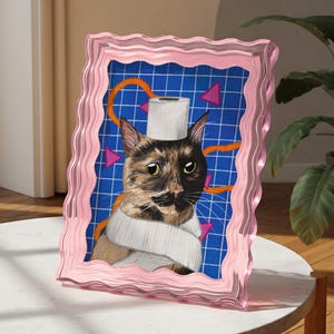 May include: A pink framed artwork depicting a cat with a toilet roll on its head and wrapped around its neck. The background is blue with a grid pattern, orange lines, and pink triangles. The frame has a wavy edge.