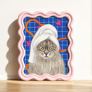 May include: A framed print of a cat wearing a white towel. The cat has grey fur and green eyes. The background is blue with a grid pattern and orange and pink geometric shapes. The frame is pink.