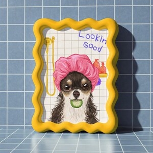 May include: A humorous illustration of a Chihuahua wearing a pink shower cap and holding a cucumber slice. The artwork is framed in a yellow, wavy-edged frame and features the text "Lookin Good". The background is a tiled wall.
