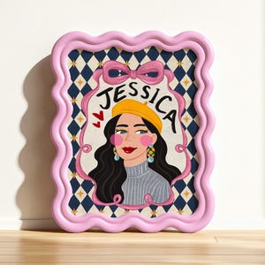 K&ouml;nnte beinhalten: Ein rechteckiges Wandbild in einem gewellten rosa Rahmen mit einer Illustration einer Person mit langem dunklen Haar, einer gelben Baskenm&uuml;tze und einem grauen Pullover. Der Name JESSICA steht in Schwarz dar&uuml;ber, umrahmt von einem rosa Band vor einem blauen Rautenmuster mit kleinen goldenen Sternen.