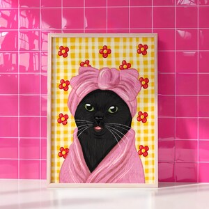 May include: A framed print of a black cat in a pink turban and robe, with green eyes and a pink tongue. The background is yellow gingham with red flower accents. The frame is light wood.