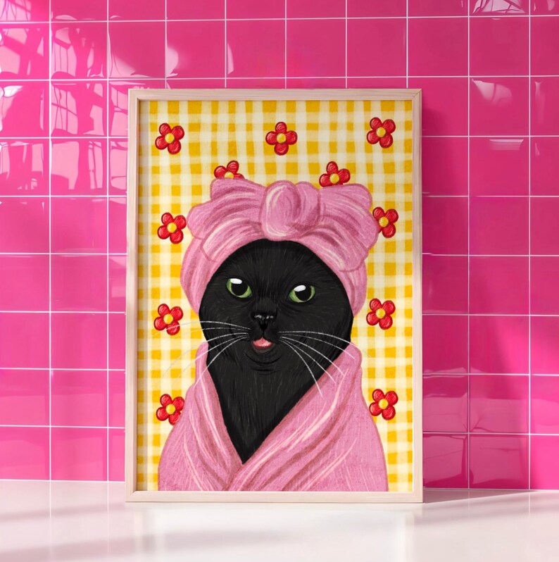 May include: A framed print of a black cat in a pink turban and robe, with green eyes and a pink tongue. The background is yellow gingham with red flower accents. The frame is light wood.