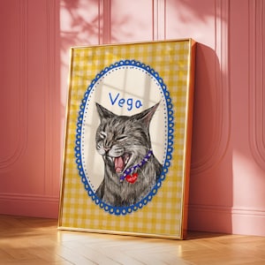 Op de afbeelding: Een ingelijst portret van een grijze tabby kat die gaapt, met een blauwe halsband met een rode hartvormige hanger. De achtergrond heeft een geel ruitjespatroon met een blauwe geschulpte rand. De naam "Vega" staat boven de kat.