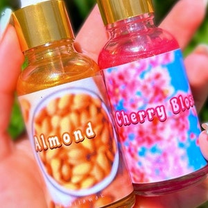 Puede incluir: Dos peque&ntilde;as botellas de vidrio con tapas y cuentagotas dorados. Una botella tiene una etiqueta con la palabra "Almond" sobre una imagen de almendras. La otra botella tiene una etiqueta con las palabras "Cherry Bloss" sobre un dise&ntilde;o floral rosa.