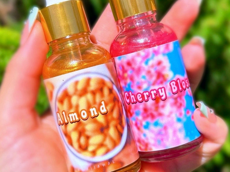 Puede incluir: Dos peque&ntilde;as botellas de vidrio con tapas y cuentagotas dorados. Una botella tiene una etiqueta con la palabra "Almond" sobre una imagen de almendras. La otra botella tiene una etiqueta con las palabras "Cherry Bloss" sobre un dise&ntilde;o floral rosa.
