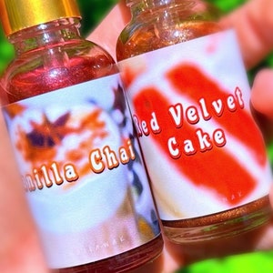 Puede incluir: Dos peque&ntilde;as botellas de vidrio con tapas doradas se sostienen en la mano. Una tiene la etiqueta "Vanilla Chai" y la otra "Red Velvet Cake". Las botellas contienen un l&iacute;quido brillante de color marr&oacute;n rojizo.