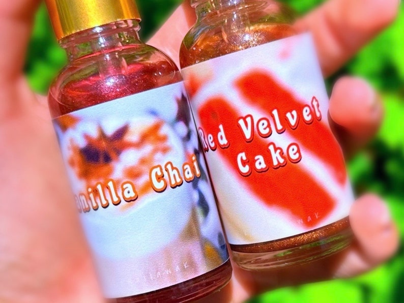 Puede incluir: Dos peque&ntilde;as botellas de vidrio con tapas doradas se sostienen en la mano. Una tiene la etiqueta "Vanilla Chai" y la otra "Red Velvet Cake". Las botellas contienen un l&iacute;quido brillante de color marr&oacute;n rojizo.