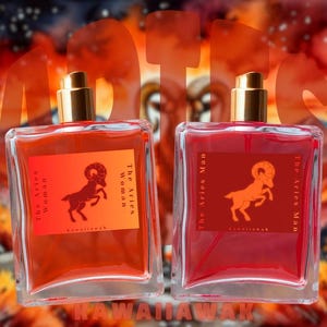 Op de afbeelding: Twee vierkante parfumflesjes met gouden doppen. E&eacute;n heeft een oranje label met een ramillustratie en de tekst "The Aries Woman". De andere heeft een rood label met een ramillustratie en de tekst "The Aries Man".