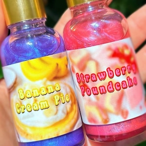 Puede incluir: Dos botellas peque&ntilde;as con tapas doradas, una llena de l&iacute;quido morado y la otra de l&iacute;quido rosa. La botella morada tiene una etiqueta que dice "Banana Cream Pie". La botella rosa tiene una etiqueta que dice "Strawberry Poundcake".