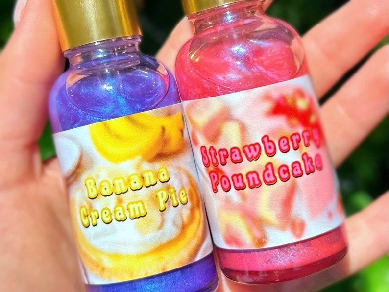 Puede incluir: Dos botellas peque&ntilde;as con tapas doradas, una llena de l&iacute;quido morado y la otra de l&iacute;quido rosa. La botella morada tiene una etiqueta que dice "Banana Cream Pie". La botella rosa tiene una etiqueta que dice "Strawberry Poundcake".