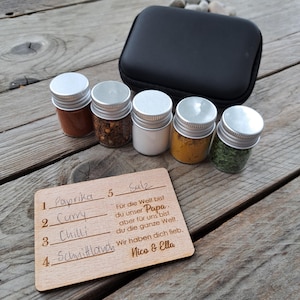 Può includere: Un set di spezie con cinque piccoli barattoli di vetro con coperchi argentati, riempiti con varie spezie, e una custodia nera. Una scheda di legno elenca le spezie e un messaggio in tedesco. Le spezie includono paprika, curry, peperoncino e sale.