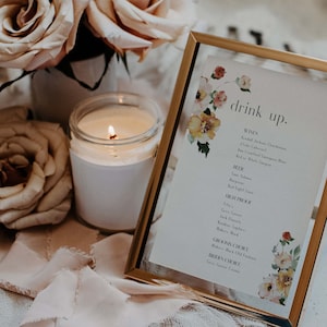 May include: A gold framed sign with a floral design and the text "drink up." The sign lists different wine, beer, and cocktail options. The sign is sitting on a white surface with a lit candle and pink roses.