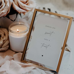 May include: A gold-framed sign with the text "babies are sweet take a treat" and a small heart symbol. The sign is sitting on a white surface with a lit candle and a bouquet of beige roses.