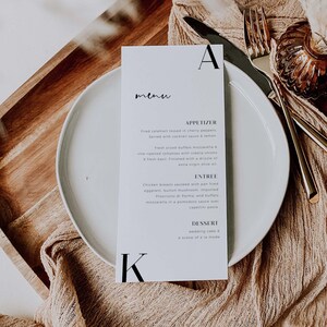 Peut inclure: Une carte de menu blanche avec du texte noir sur un plateau en bois avec une assiette blanche, une serviette en tissu marron et des couverts en or. Le menu comprend des entrées, des plats principaux et des desserts. Le texte sur la carte de menu se lit comme suit : "menu", "APPETIZER", "Fried calamart tossed in cherry peppers Served with cocktail sauce & lemon.", "Fresh sliced buffalo mozzarella & vine ripened tomatoes with vidalia onions & fresh basil Finished with a drizzle of extra virgin olive oil", "ENTREE", "Chicken breasts sauteed with pan fried eggplant, button mushroom, imported Prosciutto di Parma, and buffalo mozzarella in a pomodoro sauce over capellini pasta", "DESSERT", "wedding cake & a scoop of a la mode"