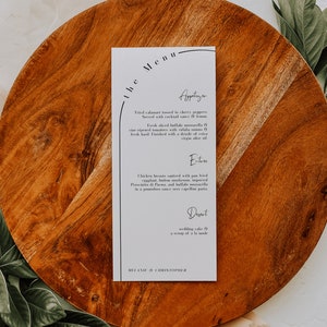 May include: A white menu card with a black border and a curved line at the top. The menu is titled "The Menu" and lists appetizers, entrees, and dessert. The appetizers include fried calamari tossed in cherry peppers, served with cocktail sauce and lemon, and fresh sliced buffalo mozzarella with vine-ripened tomatoes, vidalia onions, and fresh basil. The entrees include chicken breasts sauteed with pan-fried eggplant, button mushrooms, imported prosciutto di Parma, and buffalo mozzarella in a pomodoro sauce over capellini pasta. The dessert is wedding cake with a scoop of a la mode. The menu is signed "Melanie & Christopher."