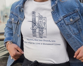 Può includere: T-shirt bianca con un'illustrazione blu navy di libri e motivi floreali. Il testo recita: "books, she has found, are the way to live a thousand lives". La persona indossa una giacca di jeans.