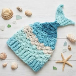 May include: A crocheted mermaid tail hat in shades of blue and cream, with a textured scale pattern. The hat is surrounded by seashells, sea glass, and a starfish on a white wooden surface.