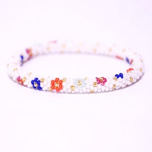 Puede incluir: Pulsera de cuentas blancas con detalles de flores de colores. La pulsera está hecha de pequeñas cuentas blancas con pequeñas cuentas rojas, azules y verdes que forman flores.