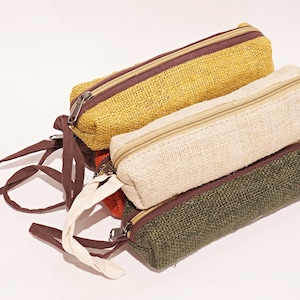 Pode incluir: Três estojos retangulares em amarelo mostarda, natural e verde oliva. Cada estojo tem um zíper correspondente e uma borda marrom contrastante. Os estojos são feitos de tecido tecido e têm uma pequena alça.