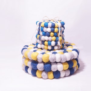 Könnte beinhalten: Ein Stapel runder Filzuntersetzer in Blau, Gelb, Weiß und Grau. Die Untersetzer bestehen aus Pompons und sind mit Schnur zusammengebunden.