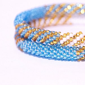 May include: Close-up of a beaded bracelet with a spiral design. The bracelet features a combination of light blue, gold, and white beads. The beads are small and tightly woven together, creating a textured surface. The bracelet is likely handmade.