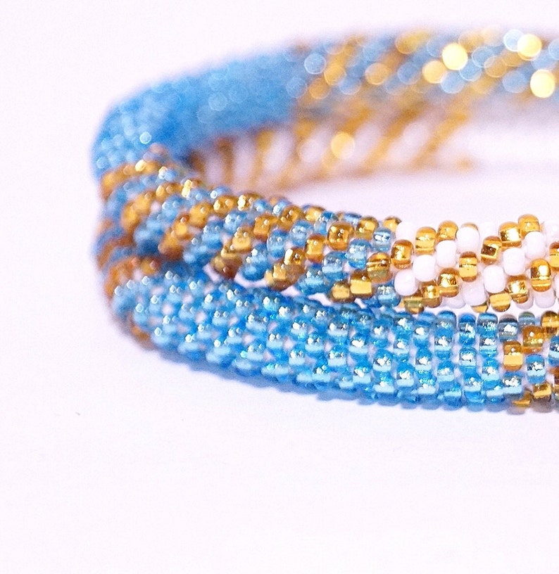 May include: Close-up of a beaded bracelet with a spiral design. The bracelet features a combination of light blue, gold, and white beads. The beads are small and tightly woven together, creating a textured surface. The bracelet is likely handmade.