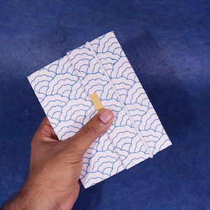 Puede incluir: Un cuaderno blanco con un diseño de ondas azul y blanco. El cuaderno tiene un cierre de cinta dorada.