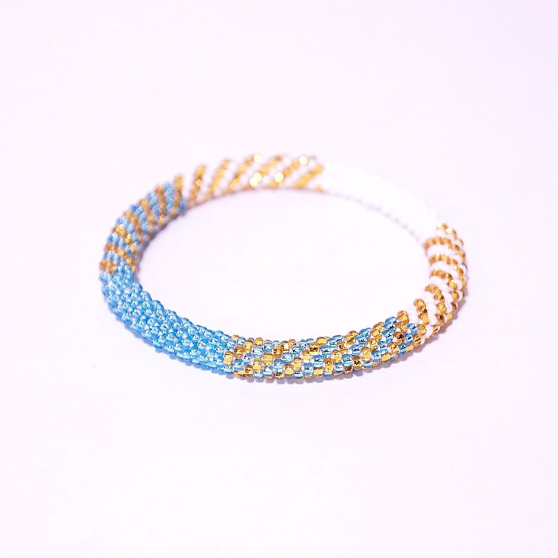 May include: A beaded bracelet with a spiral design. The bracelet features alternating sections of light blue, gold, and white beads. The beads are small and tightly woven together, creating a textured surface. The bracelet is circular and appears to be handmade.