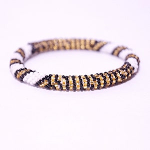 Puede incluir: Un brazalete de cuentas negras, blancas y doradas. El brazalete está hecho de pequeñas cuentas redondas que están tejidas juntas en un patrón en espiral.