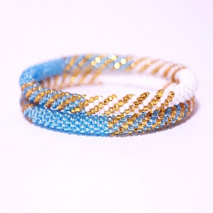 May include: Two beaded bracelets. One bracelet is light blue, the other is white, and both have gold beads. The bracelets are made of small, round beads woven together to form a spiral pattern. The bracelets are stacked on top of each other.