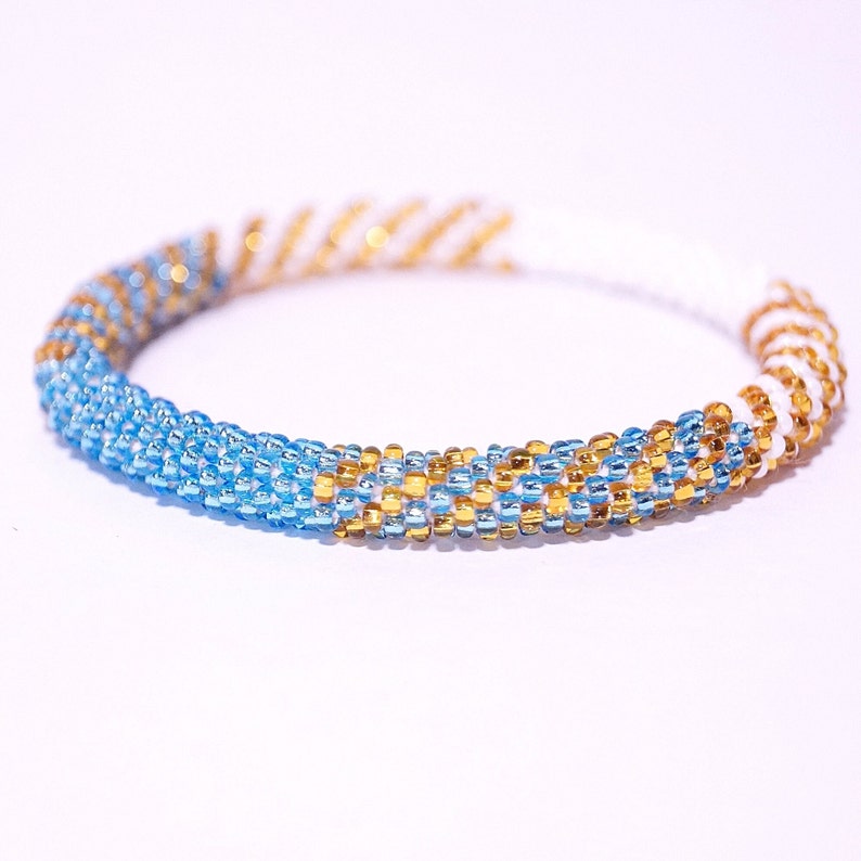 May include: A beaded bracelet featuring a spiral design. The bracelet is primarily light blue with gold and white accents. The beads are small and tightly woven, creating a textured surface. The bracelet is a circular shape.
